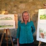 Female guest standing next to two easels showing watercolour paintings of Tuscan landscape.
