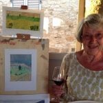 Mature lady with glass of wine next to easel and watercolour painting.